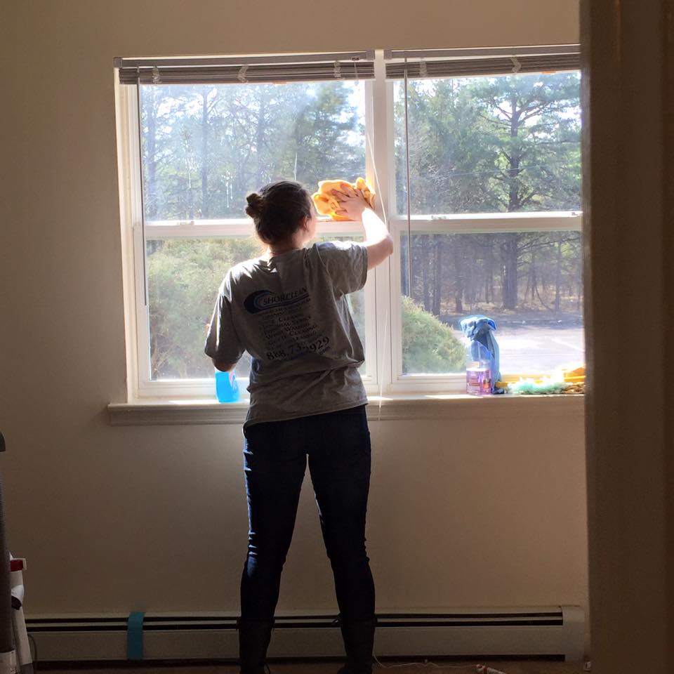 A person in a gray t-shirt and jeans cleans a window with a yellow cloth inside a room.