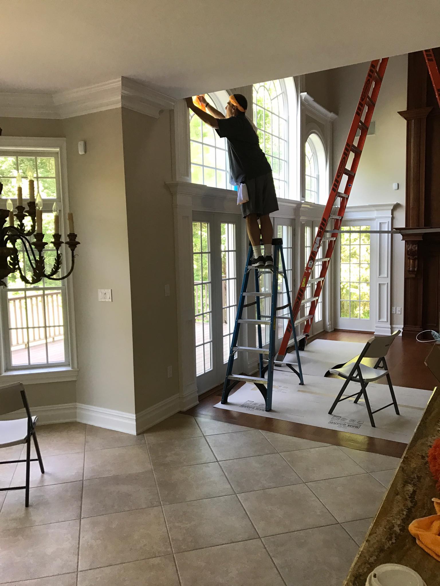 A person on a tall ladder reaches up to clean or work on the top section of large, floor-to-ceiling windows.