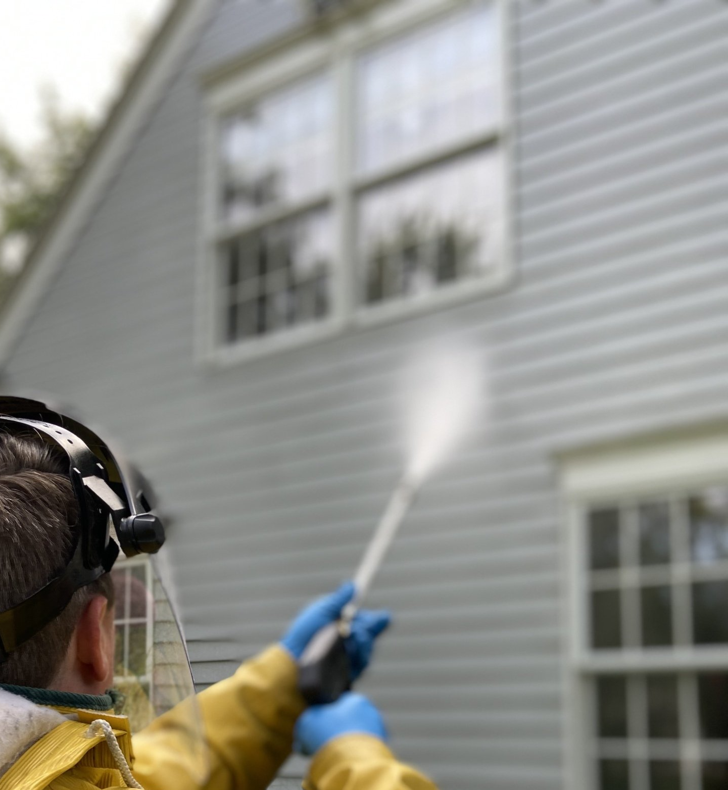 A person wearing a face shield and yellow rain jacket uses a pressure washer to clean the light-colored siding of a house.