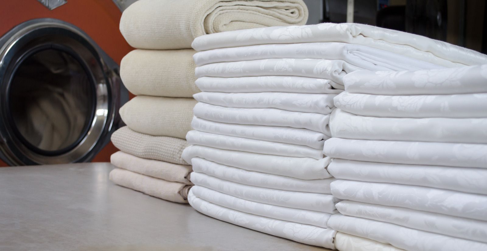 Two stacks of folded, white and cream-colored linens sit on a table in front of a laundry machine.