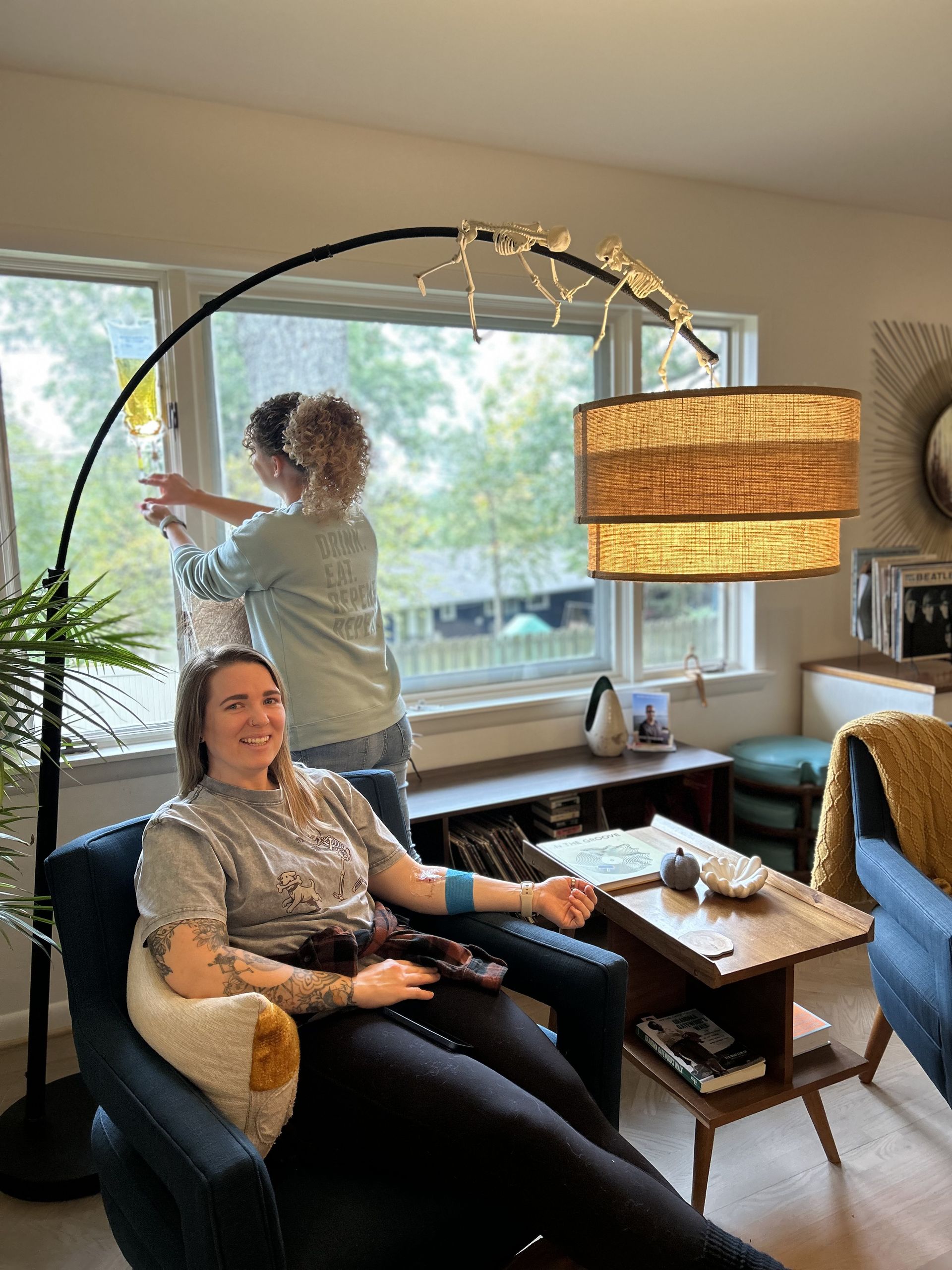 Woman in armchair smiles as another person adjusts a window covering. Living room with arch lamp.