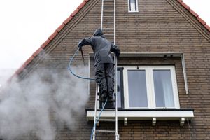 Person in waterproof suit on ladder, cleaning brick house with pressure washer, steam visible.