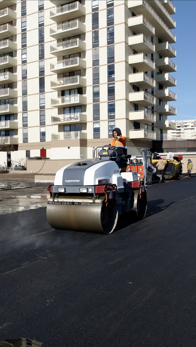 A man is driving a roller on a road in front of a building.