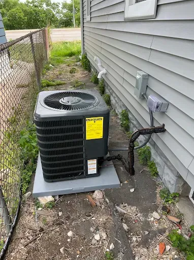 Air conditioning unit next to a gray house, on a gray platform, next to a chain-link fence.