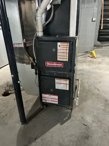 Goodman furnace in a basement setting, black casing with red logo and warning labels.
