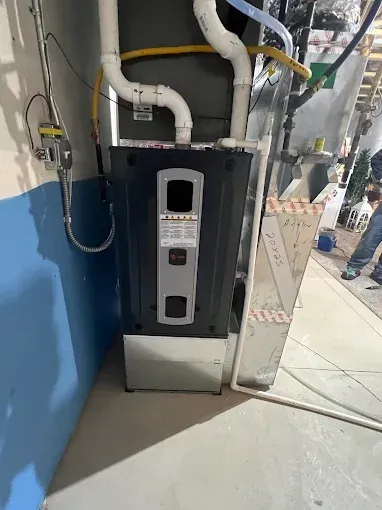A furnace installed in a basement, with white and gray pipes, next to a blue and gray wall.