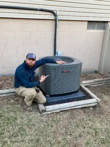 A man kneels next to an AC unit, pointing at it. He's wearing work clothes, outdoors.