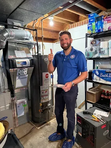 Man in blue shirt, thumbs up next to water heater in a basement.