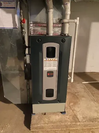 Dark gray Trane furnace in a basement setting, with ductwork and a label visible.