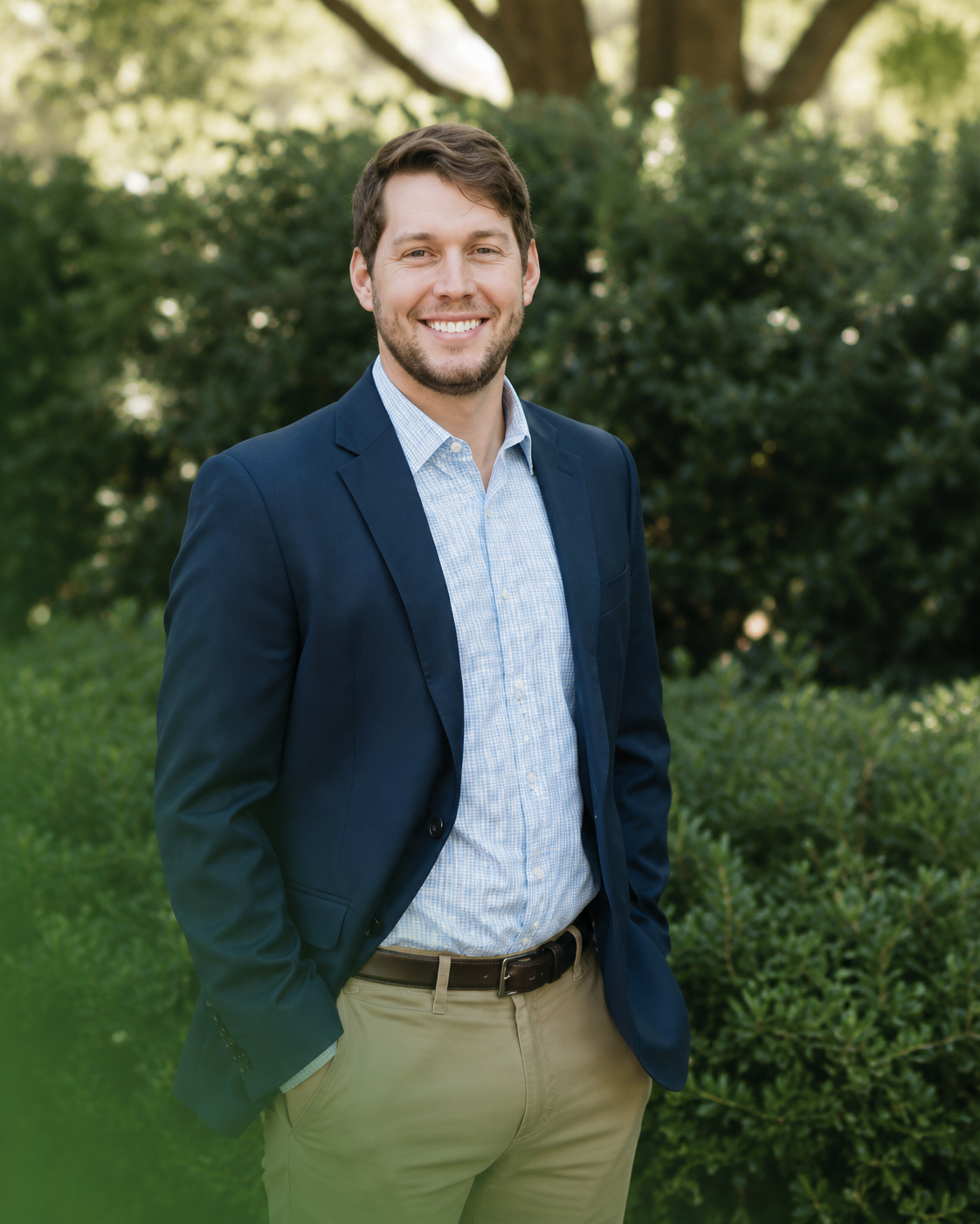 Smiling man in a navy blazer and khaki pants standing outdoors in a green garden.