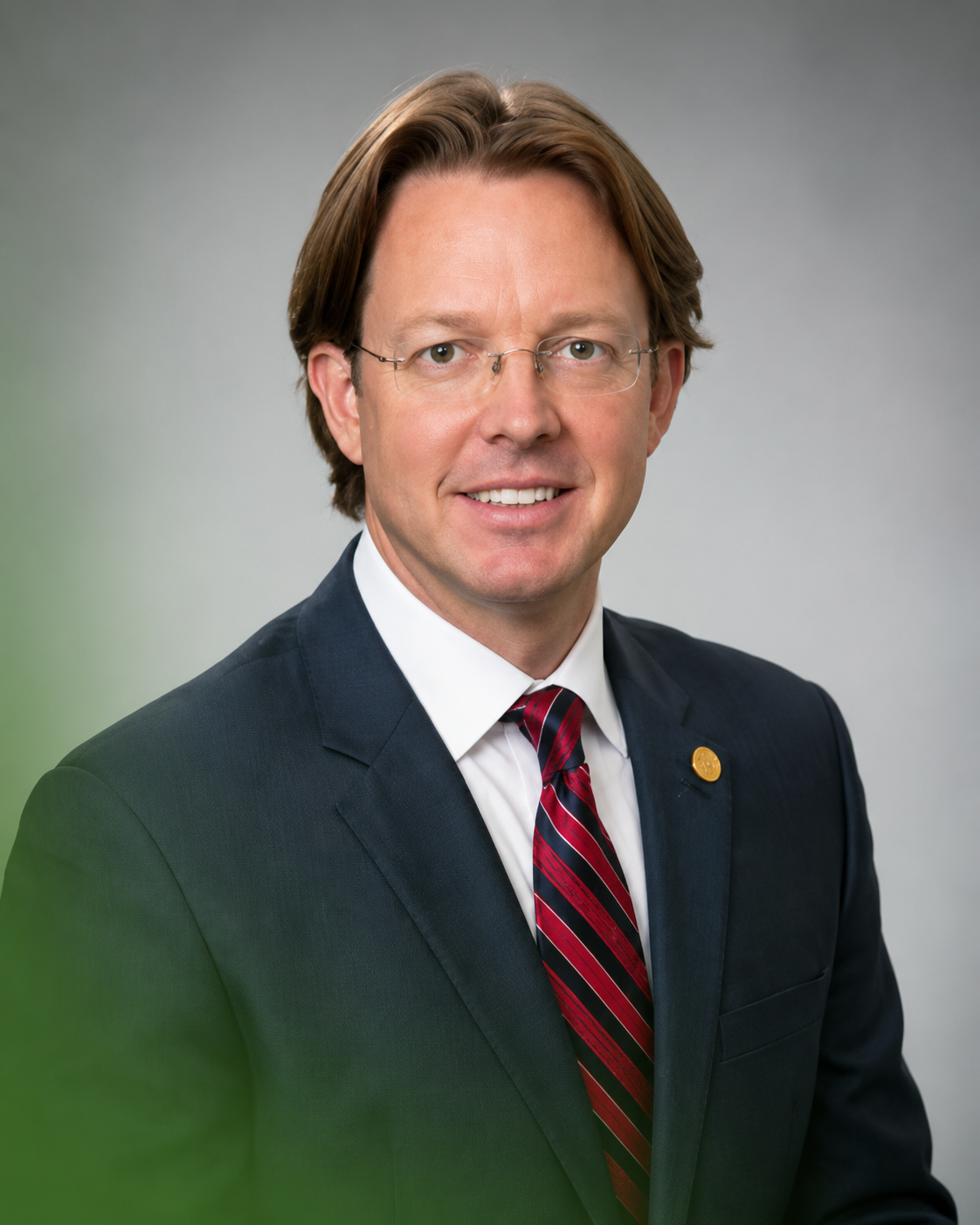 Portrait of a man in a dark suit, red striped tie, and glasses against a gray background.