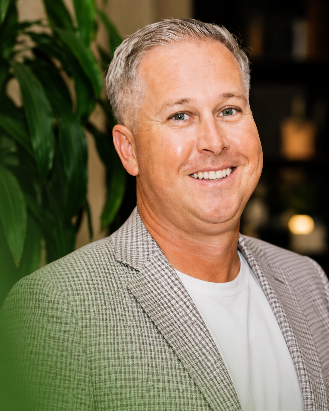 Smiling man in a light gray blazer and white shirt indoors with blurred plants in the background