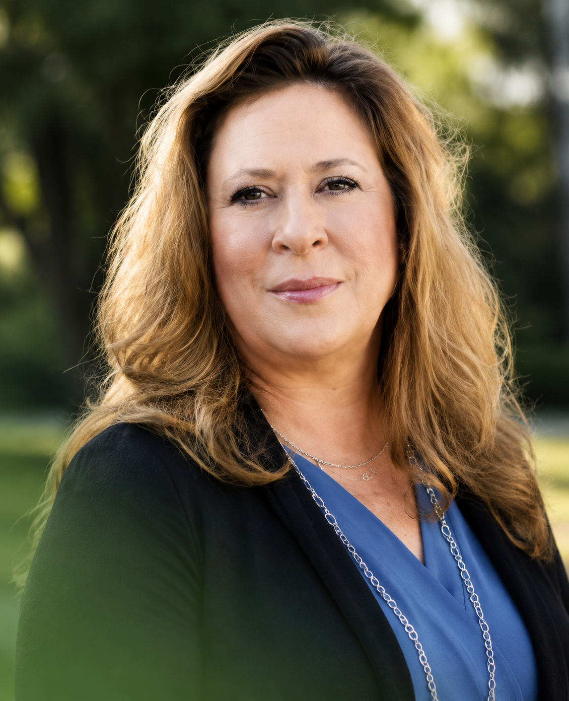 Woman standing outdoors, wearing a black blazer over a blue top and necklace, with a blurred green background.