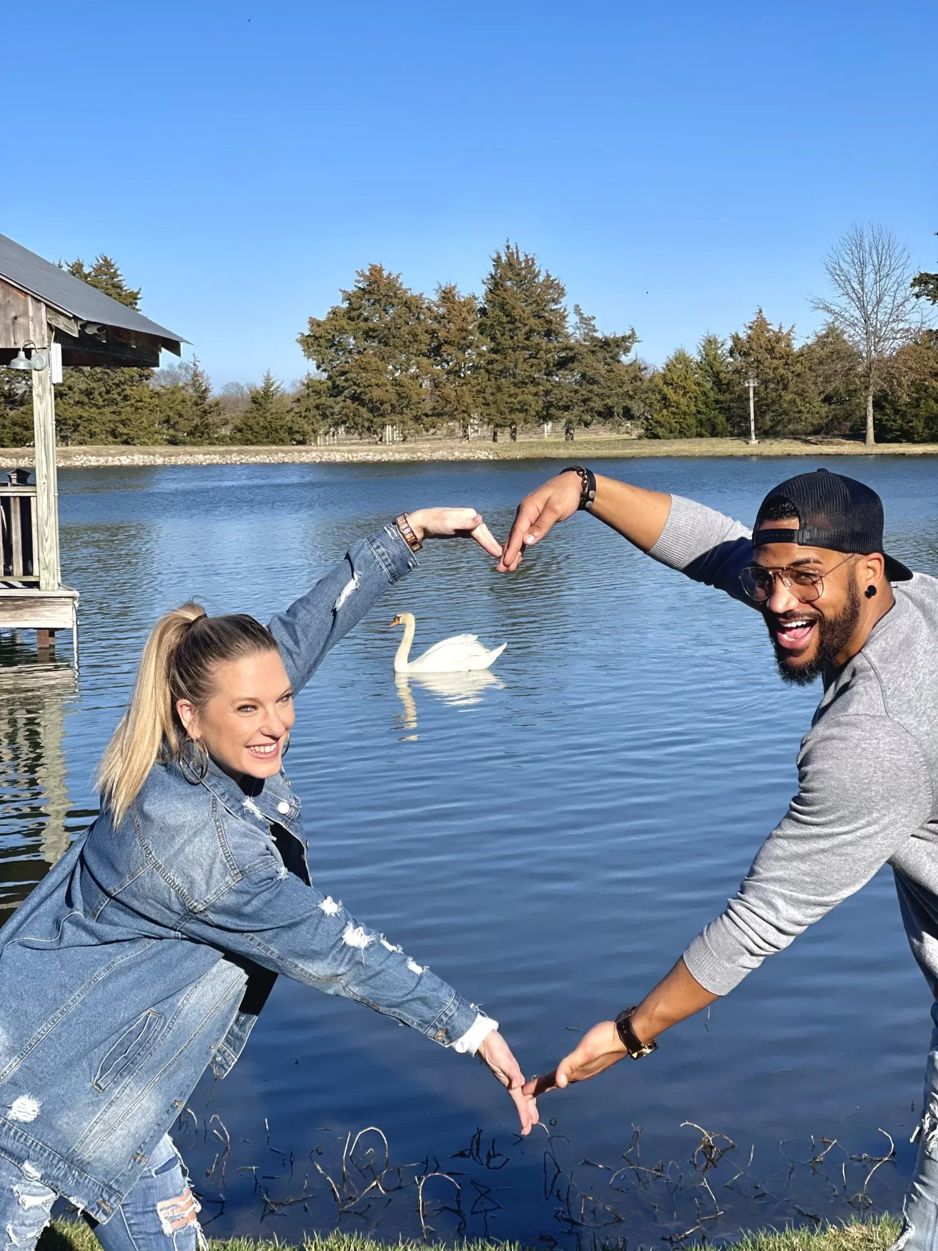 A man and a woman are making a heart shape with their hands in front of a lake.