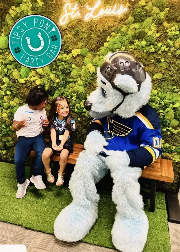 Two little girls are sitting on a bench next to a mascot.