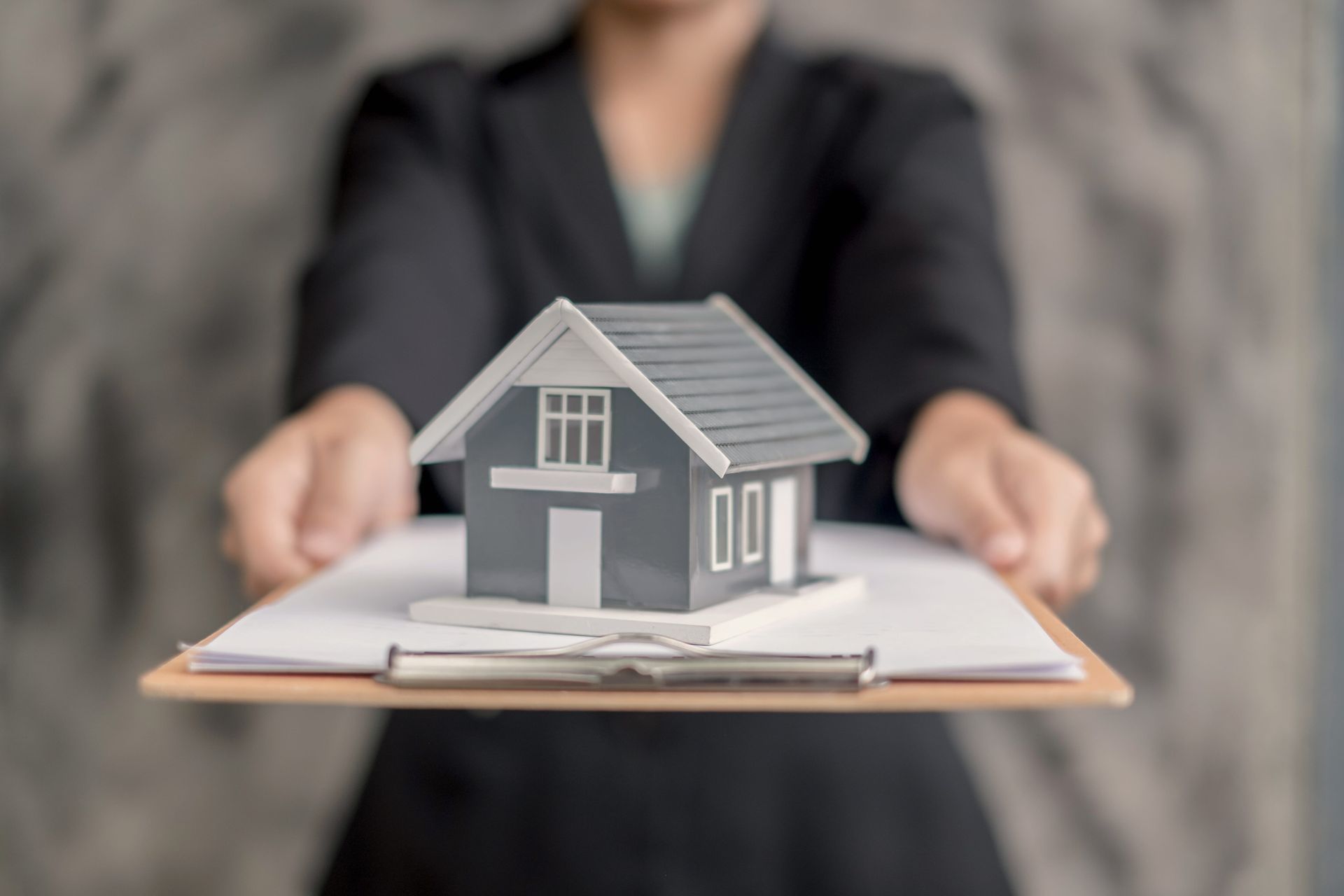 Hands holding a clipboard with a small gray model house in front of a blurred person