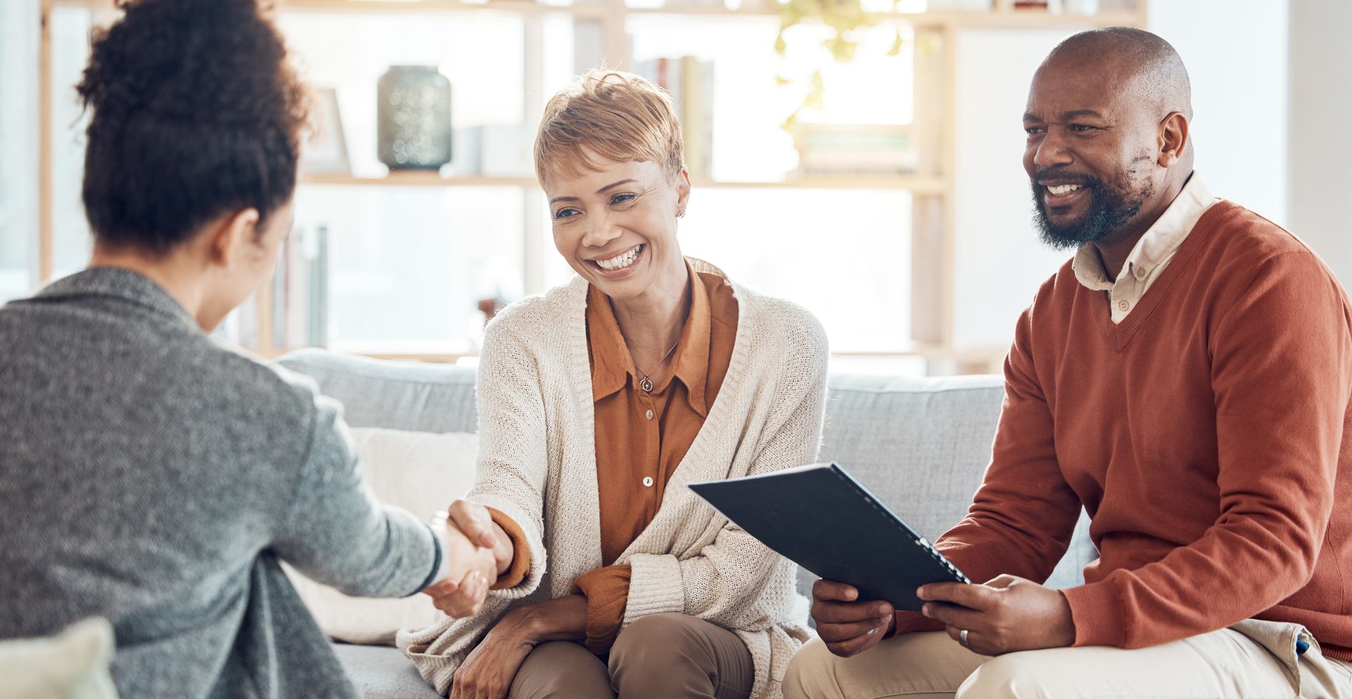 Woman shaking hands with a couple sitting on a couch; all smiling.  Light-filled room.