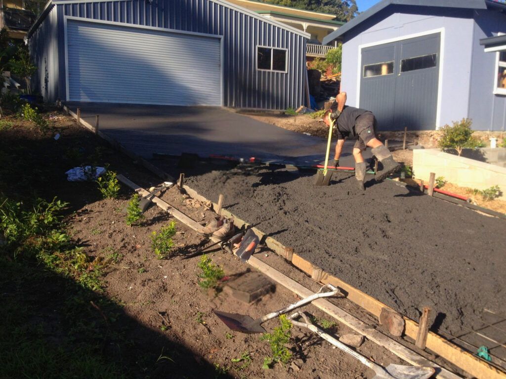 Person shoveling wet soil in a backyard beside a house and garage, with wooden forms in the foreground