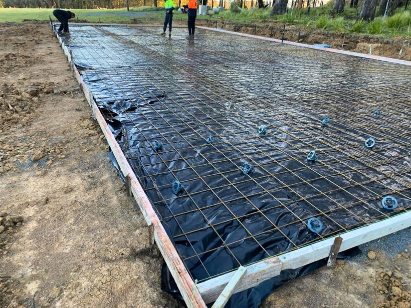 Concrete slab under construction, covered with rebar and black plastic, with workers in the background.