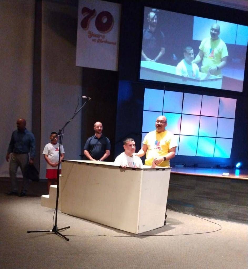 Baptism ceremony on a stage. Man in yellow shirt baptizes another in a white pool. Others watch.