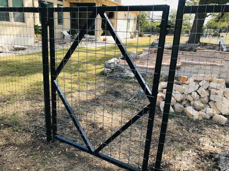 Black metal gate with diagonal bracing in a yard, beside stacked stone blocks and a wire fence.
