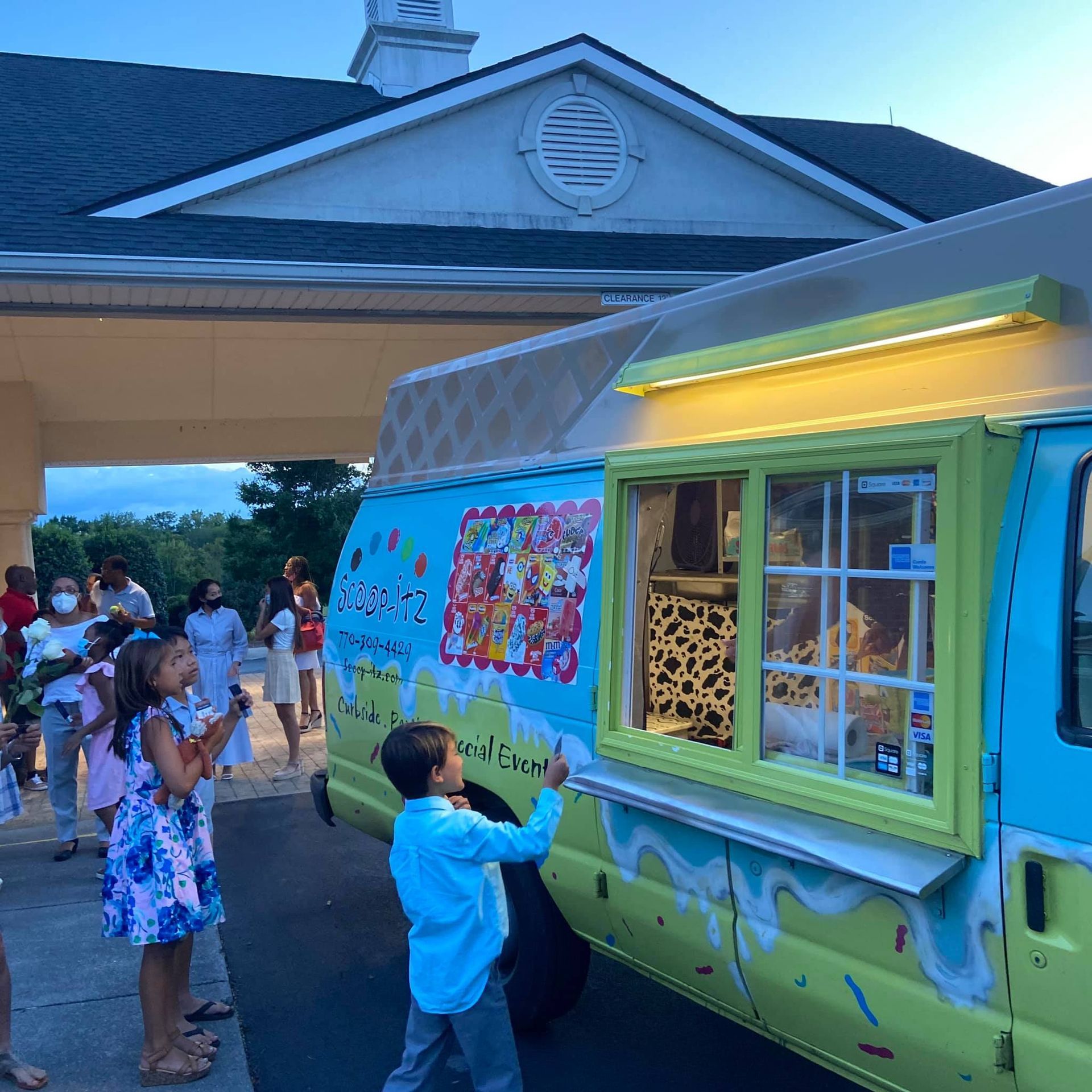 Children wait at a brightly painted blue and green ice cream truck parked under a building portico at dusk.