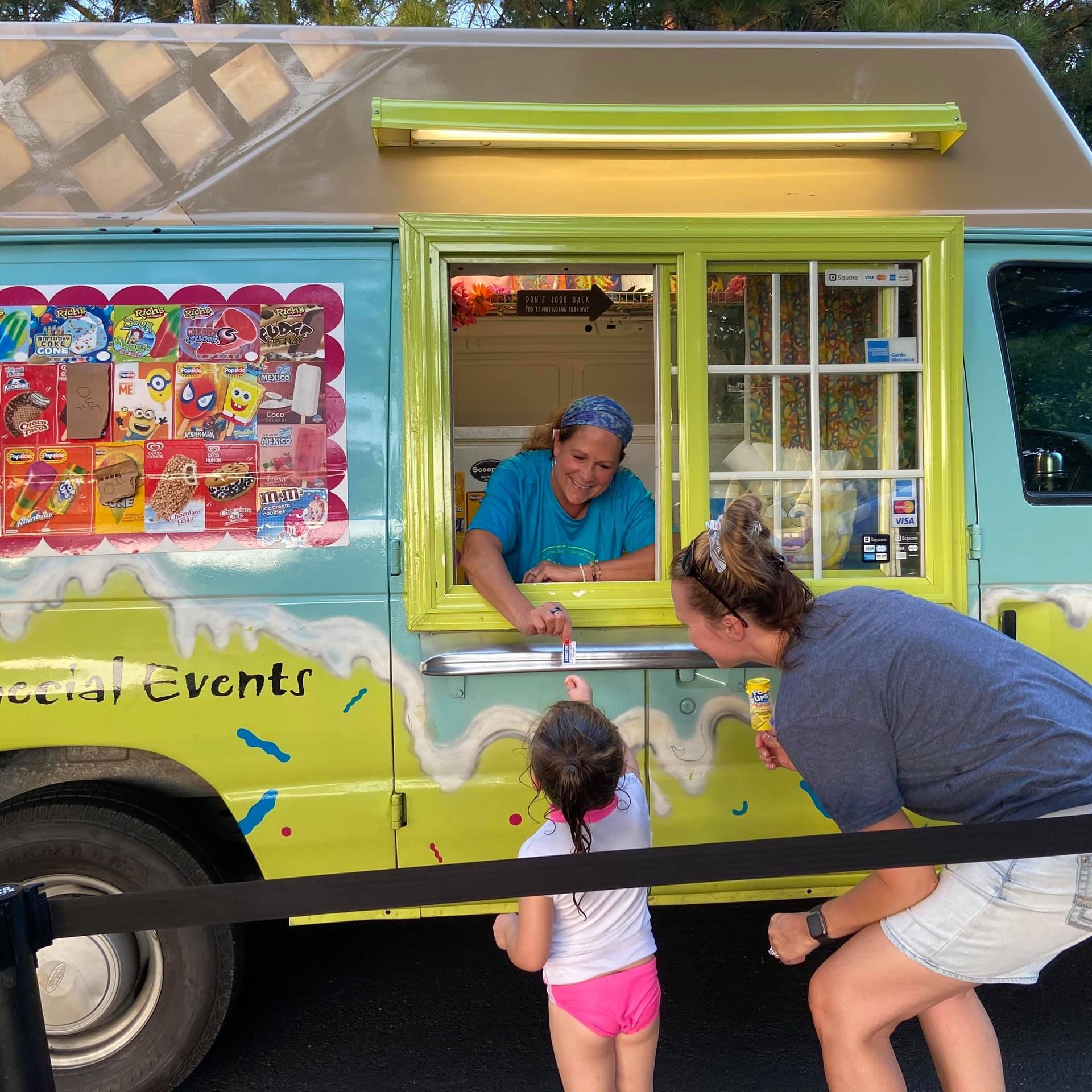 A person receives an ice cream treat from a smiling vendor inside a bright yellow and teal ice cream truck.