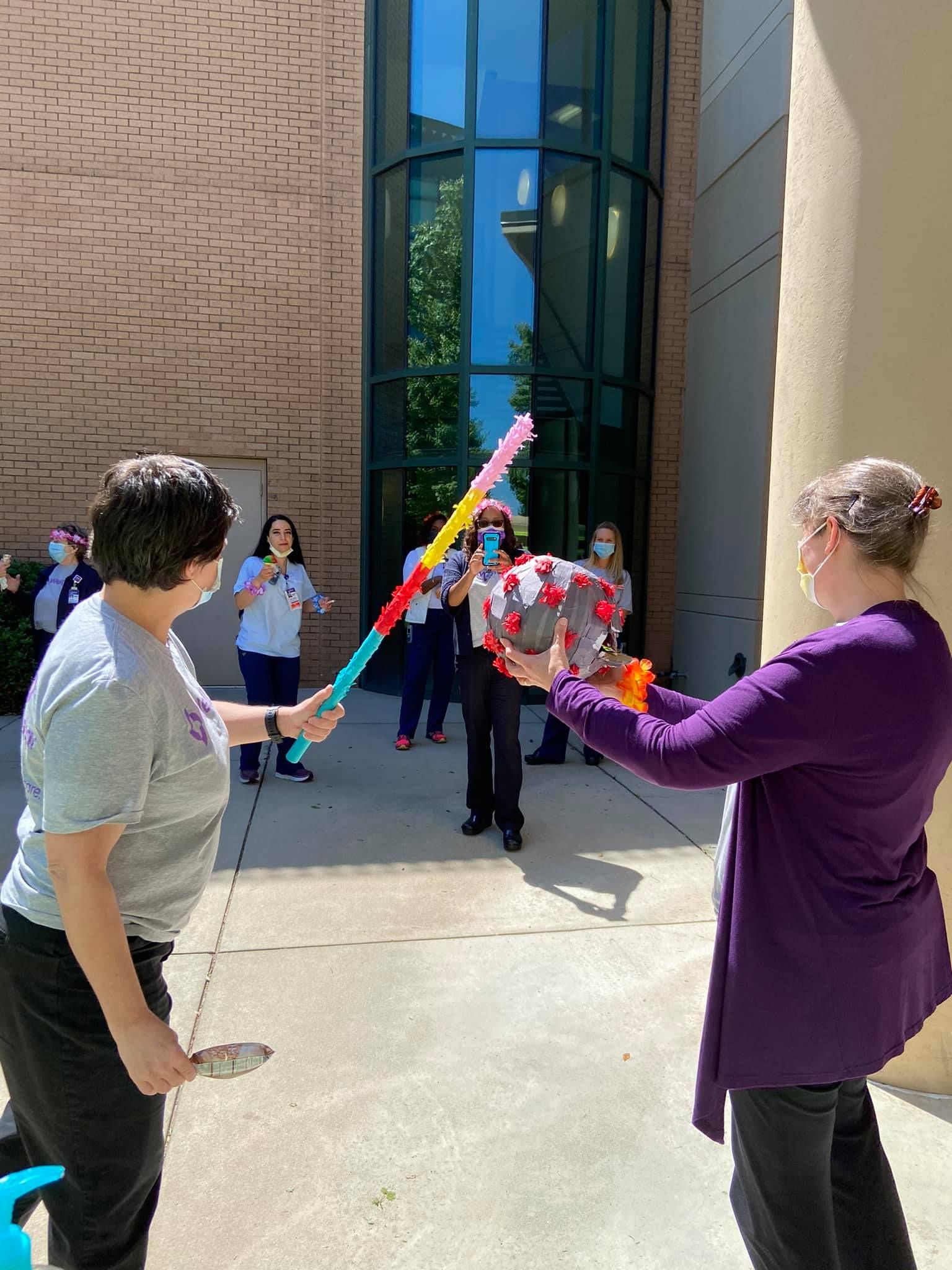A person in a gray shirt uses a long, colorful stick to strike a piñata held by another person in a purple cardigan.