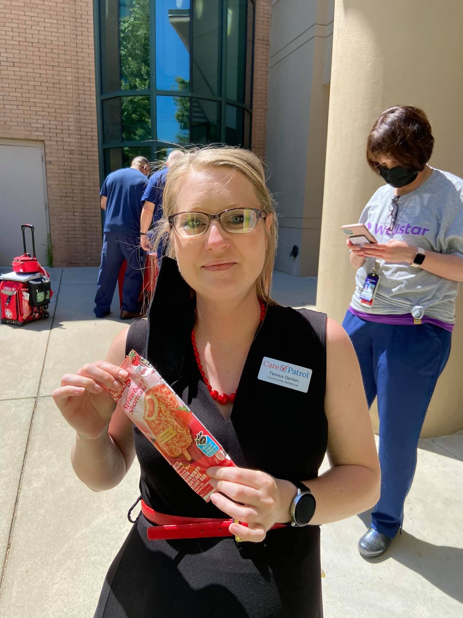 A person in a black dress holds a red packaged ice cream treat outdoors, with another person in the background.