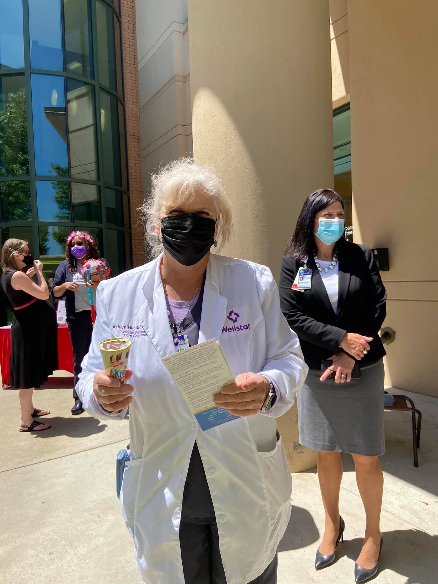 A person in a white lab coat holds a bell and card, standing outdoors near others in professional attire.