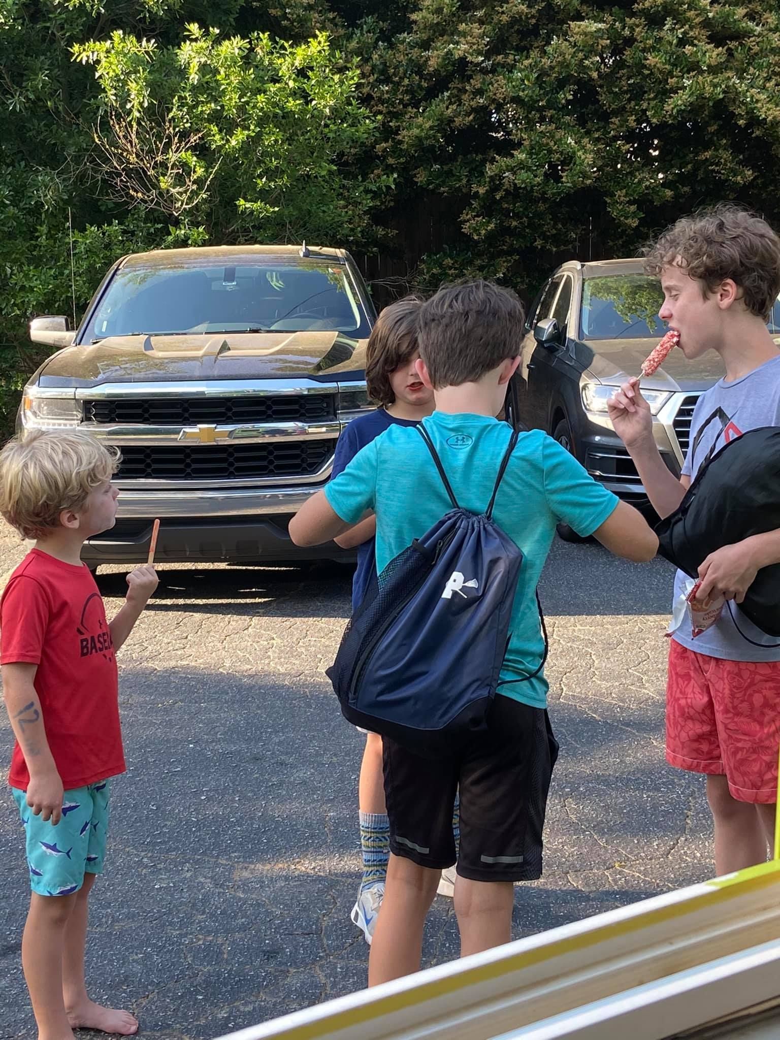 Four children gather outdoors in a parking lot, some holding ice pops, with two vehicles parked in the background.