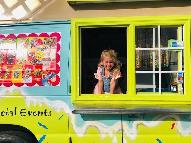 A smiling child leans out the service window of a brightly colored, lime green and blue ice cream truck.