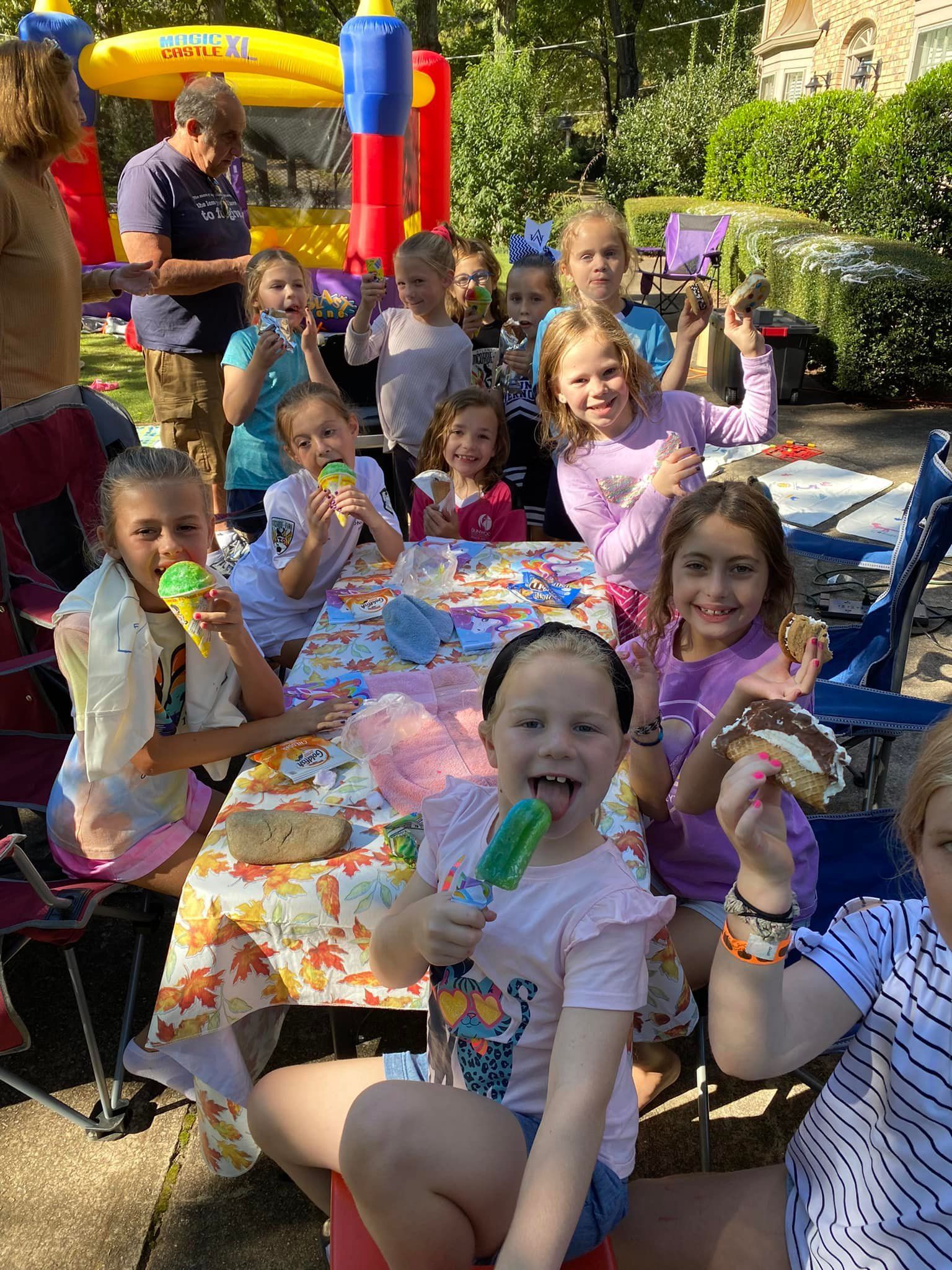 Children enjoying snacks and treats at an outdoor party with a bounce house in the background.