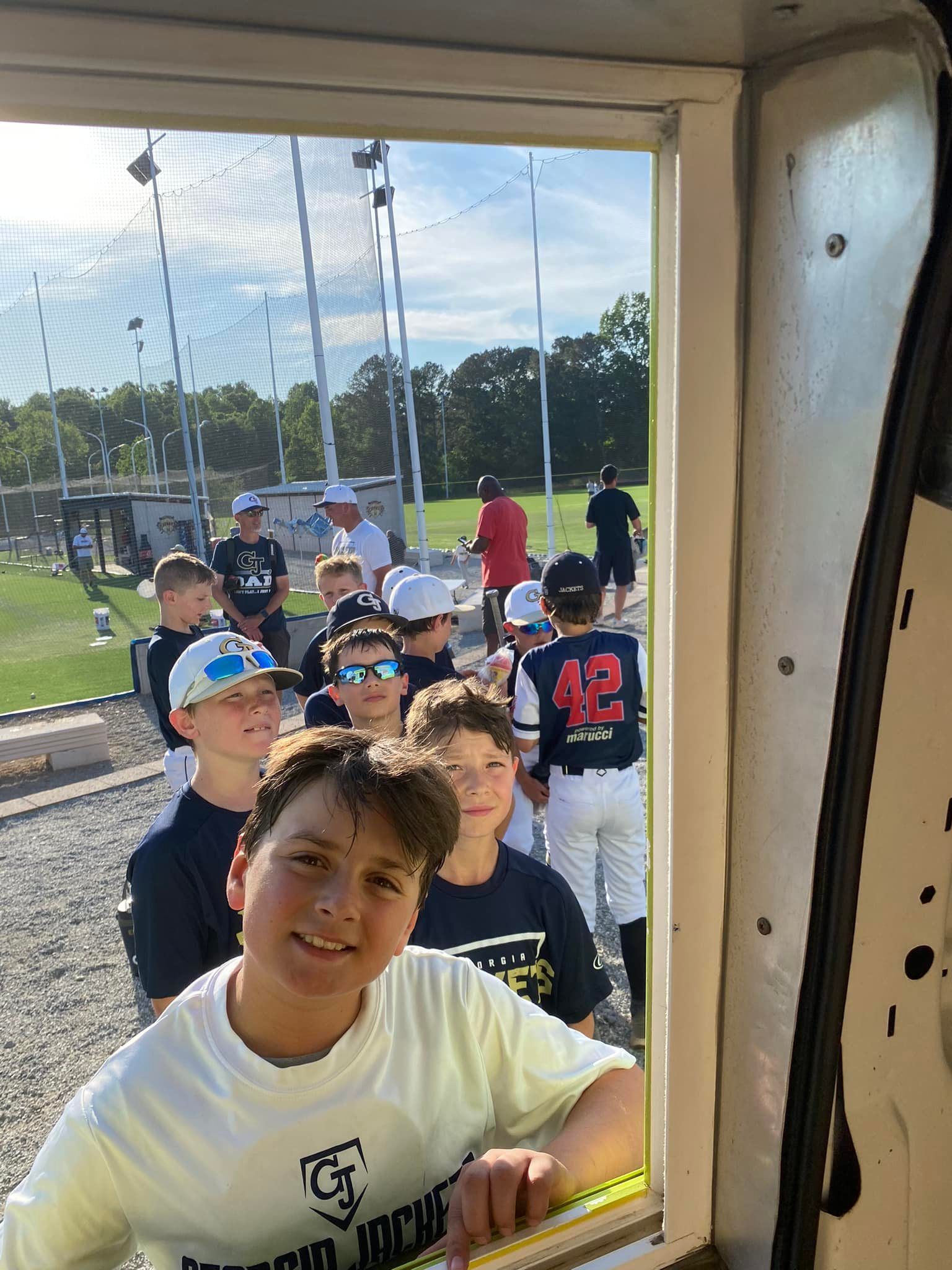 A boy smiles in the foreground, with teammates in baseball uniforms visible through a window at a sunny sports field.