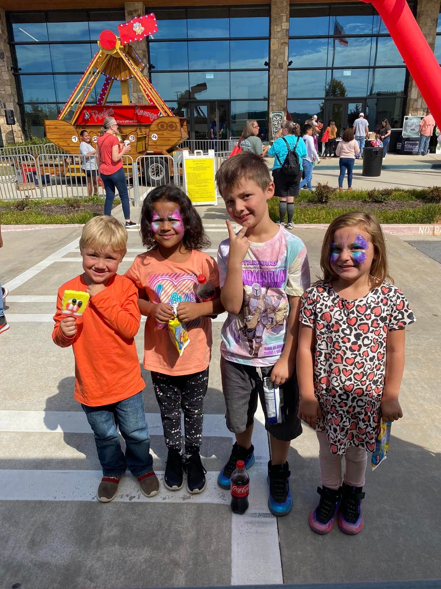 Four children stand together smiling on a sunny day at an outdoor event with a wooden ship display in the background.