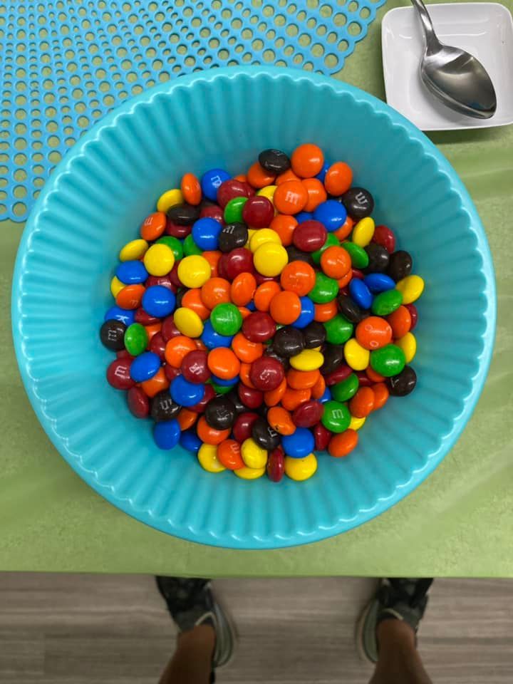 A teal, fluted bowl filled with colorful candy-coated chocolates sits on a green surface next to a spoon.