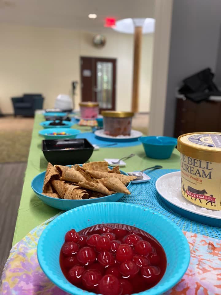 A table set up for an ice cream social, featuring bowls of cherries, waffle cones, and tubs of Blue Bell ice cream.