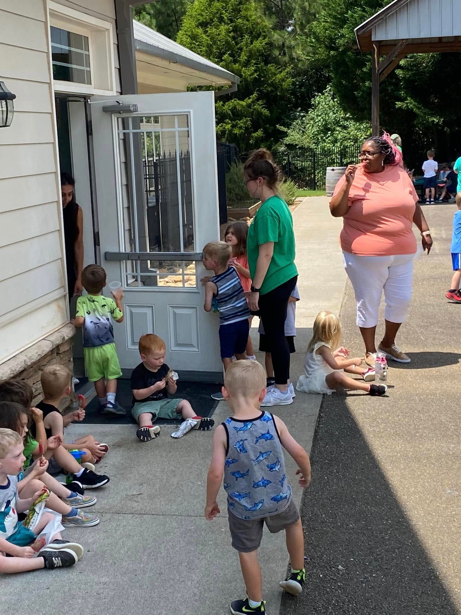 A group of children and adults gather outside an open building door on a sunny day.