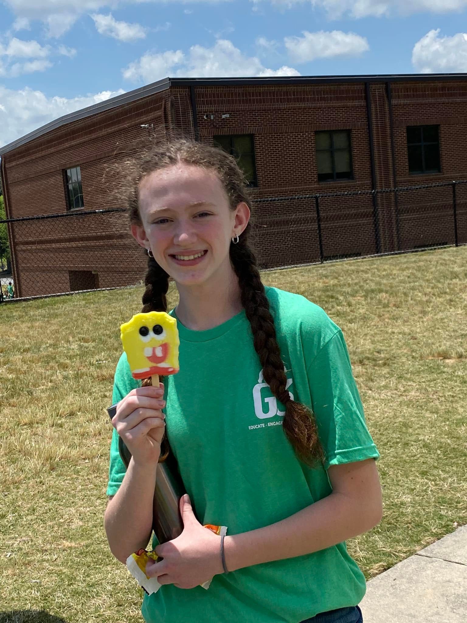 A smiling person with braided hair in a green shirt holds a Spongebob Squarepants popsicle outdoors near a brick building.