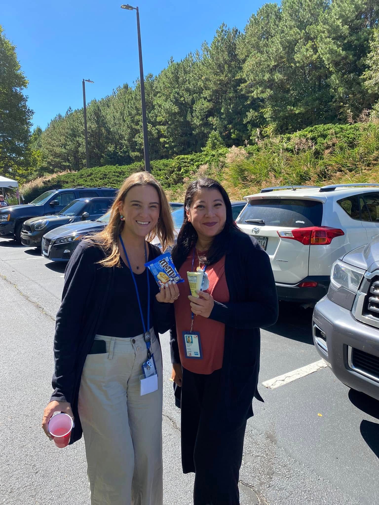 Two people stand in a parking lot on a sunny day, smiling while holding small food items and wearing ID lanyards.
