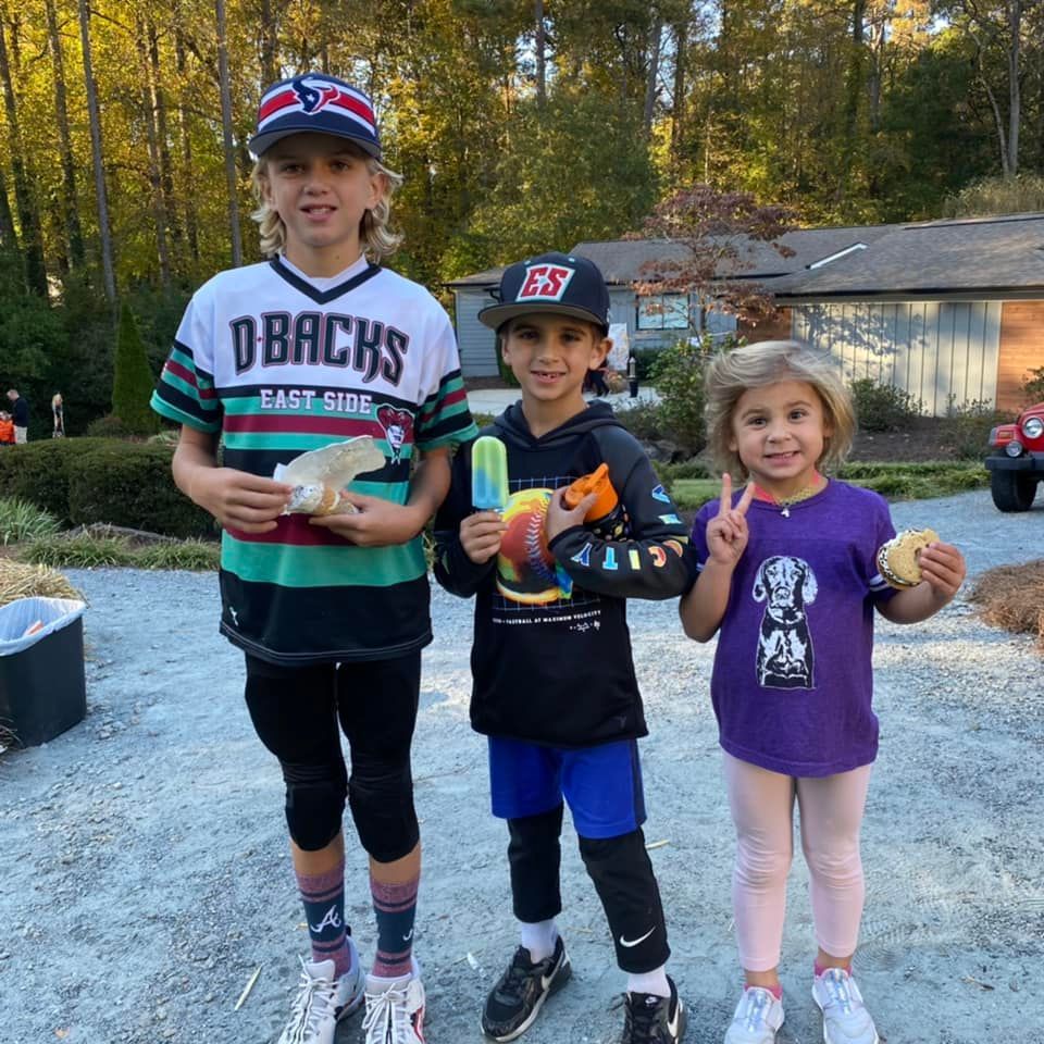 Three children in casual clothes stand outdoors holding snacks, including popsicles and cookies, smiling at the camera.