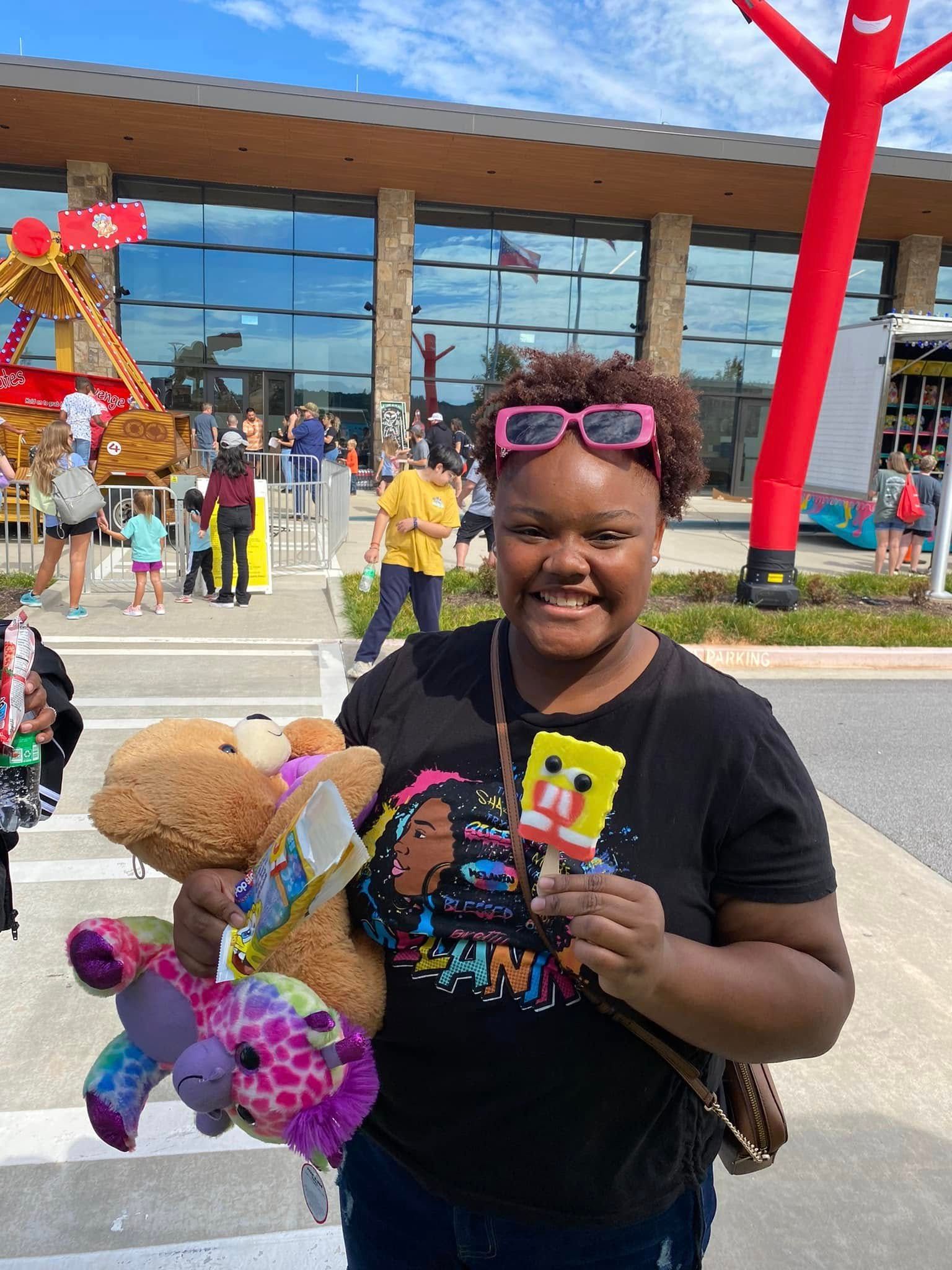 A smiling person wearing pink sunglasses holds a teddy bear, a colorful plushie, and a SpongeBob ice cream bar outdoors.