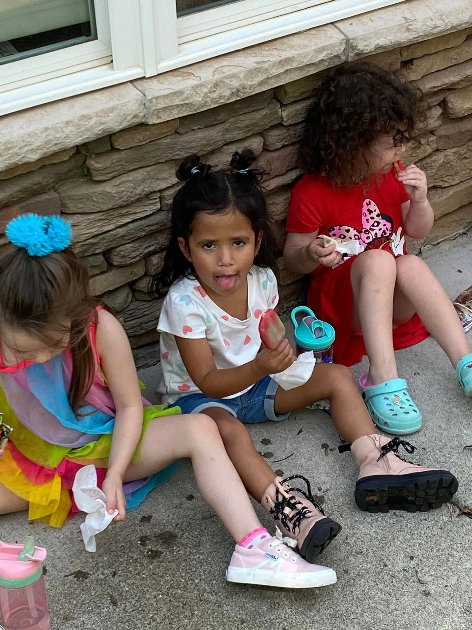 Three children sit on concrete outside against a stone wall, enjoying snacks and wearing colorful summer clothes.