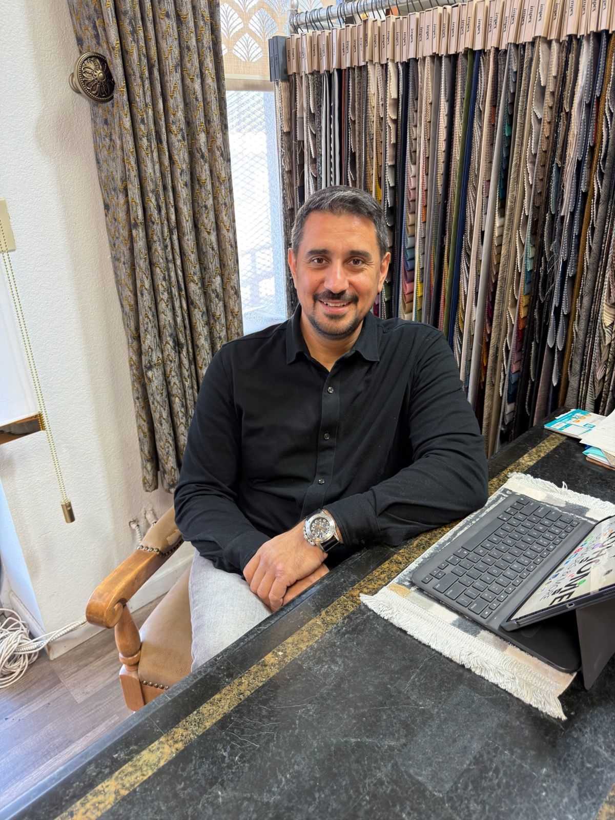 Man seated at a desk with a laptop, smiling in a office setting with curtains behind him.