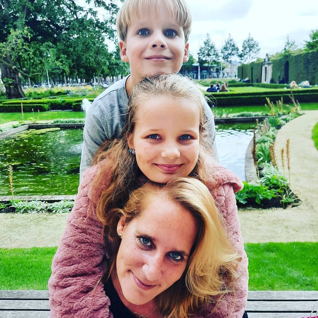 Vrouw en twee kinderen op elkaar gestapeld, lachend. Buitenomgeving: park met vijver en groen.