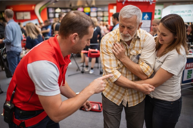 Man in rood vest wijst oudere man de weg naar zijn hand; vrouw helpt. Binnen.