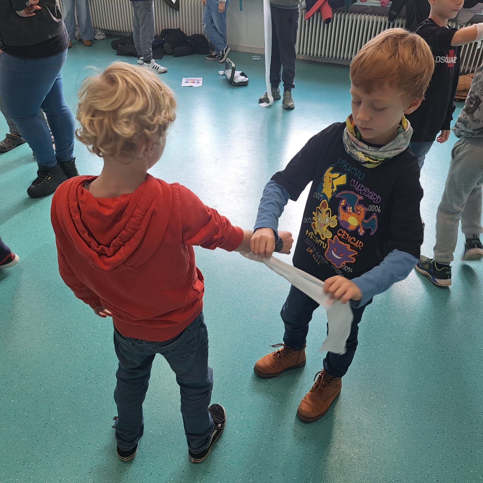 Twee kinderen trekken aan een witte strook stof. De jongen rechts draagt een zwart shirt, de ander een rode hoodie.