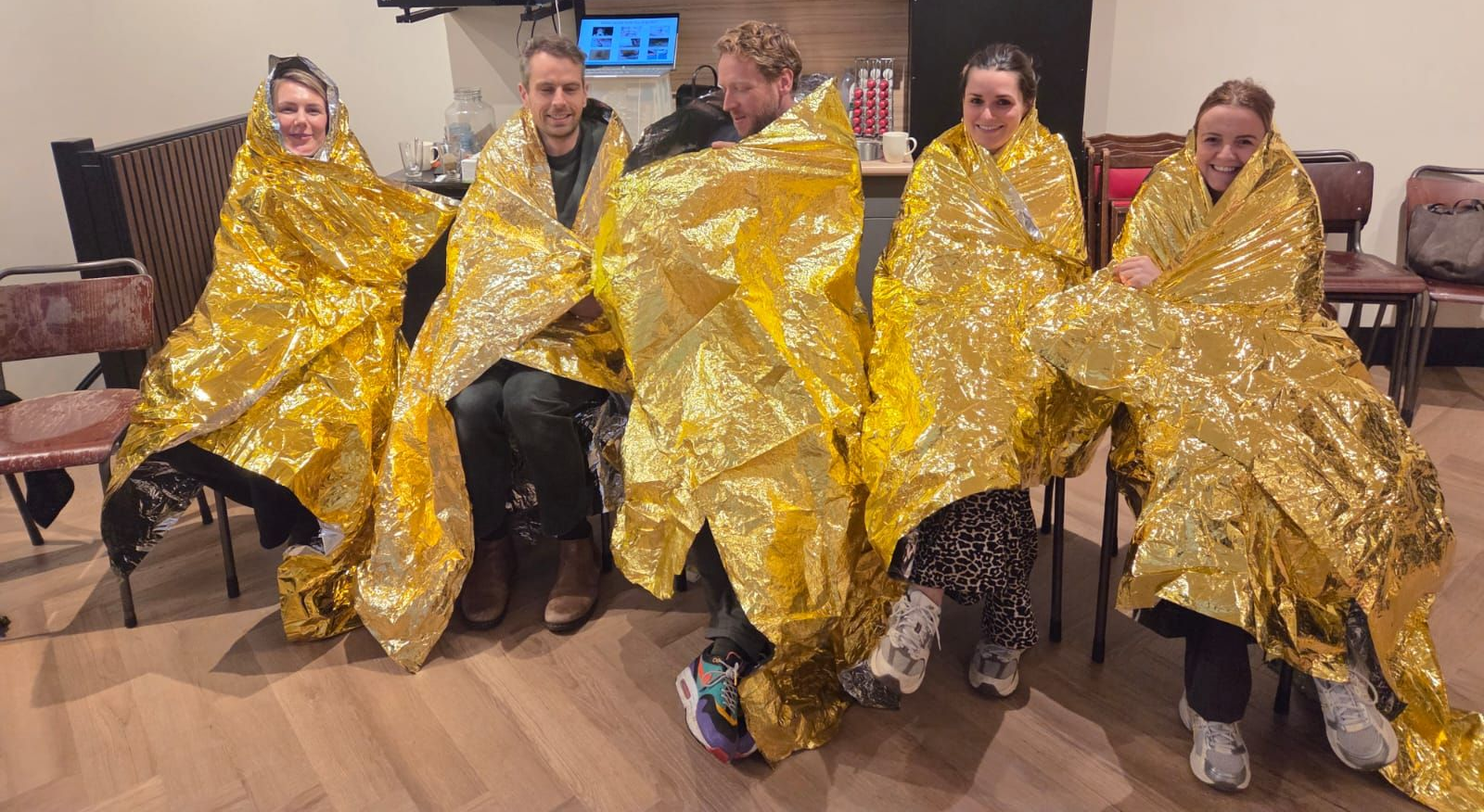 Vijf mensen, gewikkeld in dekens van goudfolie, zitten binnen, waarschijnlijk om warm te blijven.