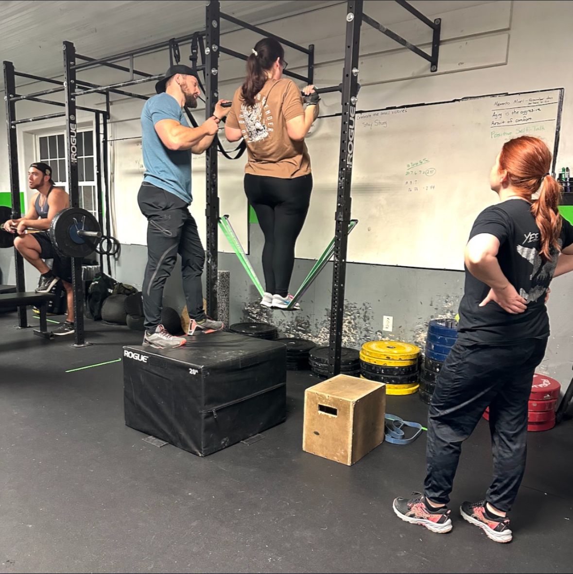 People exercising on boxes in a CrossFit gym, with a coach watching nearby.