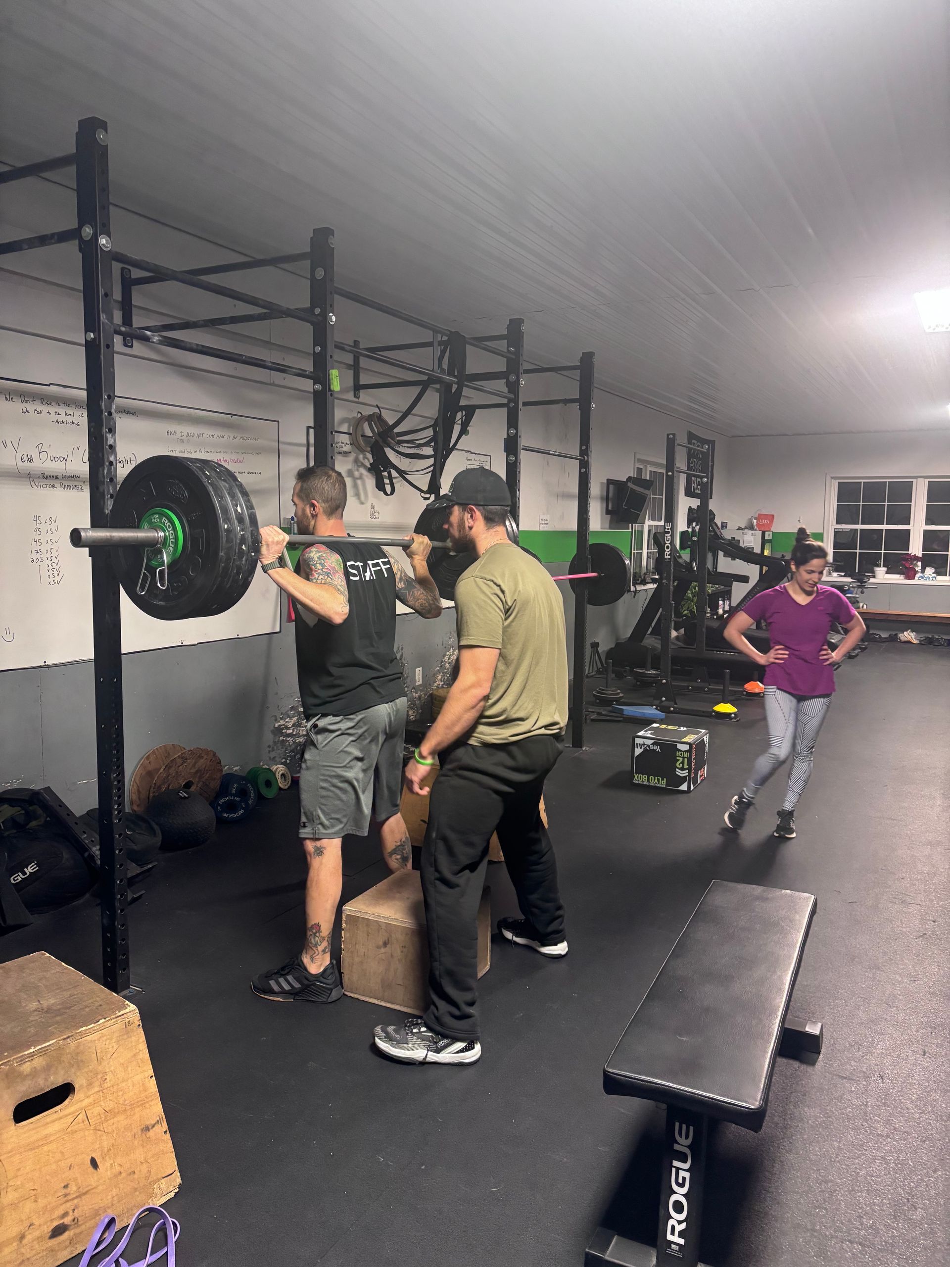 Gym class with people lifting a barbell on a squat rack, benches and equipment visible in the background.